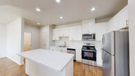 Located along N IH-35 in Austin, TX, this two-story residence offers 2 bedrooms, 3 bathrooms, and a well-organized layout spread across two floors. The main level features an open-plan kitchen and living area finished with warm hardwood-style flooring and recessed lighting throughout. The kitchen is outfitted with white shaker cabinetry, white quartz countertops, a central island, stainless steel appliances including a refrigerator, range, microwave, and dishwasher, and a double-basin sink — combining a clean, contemporary aesthetic with practical functionality. The living area flows naturally from the kitchen, creating a cohesive and bright common space. Upstairs, the spacious primary bedroom offers generous square footage with plush carpeting, a ceiling fan, and a dedicated closet. The second bedroom is also carpeted and features a window allowing natural light. Both floors are served by staircases, and the residence includes three full bathrooms — one featuring a tiled soaking tub with brushed gold fixtures, and another with a glass-enclosed walk-in shower, wood-style flooring, and a white vanity. The overall palette of whites and neutrals creates a clean, modern ambiance throughout.