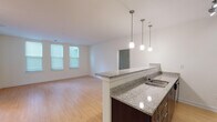 This thoughtfully designed single-floor residence offers a clean, contemporary aesthetic throughout. The open-concept layout connects the kitchen and living area seamlessly, creating a spacious and functional core. The kitchen is outfitted with dark espresso cabinetry, granite countertops, stainless steel appliances including a refrigerator, range, and built-in microwave, and a track lighting system. A peninsula with pendant lighting above provides additional prep space and serves as a natural divider between the kitchen and the expansive living area. The living space features warm hardwood flooring and large windows that invite generous natural light. The bedroom offers plush carpeting and dual windows with blinds, providing both comfort and brightness. Two bathrooms are present: one features a tub-shower combination with large-format tile surround, and both share a consistent design language of dark vanity cabinetry, white countertops, and light tile flooring. Additional features include a walk-in closet and track lighting accents. The overall color palette of whites and warm neutrals, paired with dark cabinetry contrasts, lends the space a modern yet welcoming ambiance throughout.