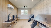 Located at 3272 State Highway 30 in Huntsville, Texas, this purpose-built dog grooming studio offers a clean, modern environment designed with both functionality and style in mind. The single-room space makes efficient use of its compact footprint, delivering a fully equipped grooming suite with a thoughtfully curated aesthetic.

The interior is defined by crisp white grid-pattern tile walls that line the majority of the space, complemented by warm wood-paneled accent walls that add contrast and a welcoming touch. A polished concrete floor with integrated floor drains ensures easy, hygienic cleanup — a practical necessity for a professional grooming environment. Recessed lighting and a sleek black ceiling fan provide comfortable, consistent illumination and climate control throughout, supported further by a wall-mounted mini-split HVAC unit.

The studio is equipped with multiple grooming tables and a stainless steel elevated bathing tub with integrated steps, allowing for efficient handling of dogs of various sizes. A glass door and a centrally positioned window flood the space with natural light and offer views of the surrounding outdoor area.

A glowing "Oh! My Dog" neon sign mounted on the wood accent wall adds a playful, branded personality to the space, rounding out a grooming studio that is as visually appealing as it is professionally appointed.