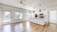 This single-floor residence is designed with a clean, contemporary aesthetic and an open-concept layout that seamlessly connects the kitchen, dining, and living areas. Light-toned hardwood-style flooring runs throughout the main living spaces, complementing the bright white walls and creating an airy, cohesive atmosphere. Multiple windows fitted with blinds allow natural light to filter in across the space.

The kitchen is a standout feature, anchored by a large center island with a built-in sink and smooth countertops. White shaker-style cabinetry lines the walls alongside a subway tile backsplash, and stainless steel appliances—including a range, microwave, and refrigerator—complete the space. Striking black pendant lights hang above the island, adding a modern contrast. A ceiling fan serves the adjoining living area.

The bedrooms offer plush neutral carpeting and ceiling fans, providing a comfortable retreat. One room includes access to an adjacent space, likely a bathroom or closet. The bathroom features white cabinetry with a stone-topped vanity, a framed mirror, and a full tub-shower combination with tile surround. A dedicated laundry area houses a full-size washer and dryer. Additional rooms with hardwood-style flooring offer flexible use, with exterior door access providing outdoor connectivity.