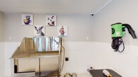Located at 1331 Wicklow Dr in Cary, NC, this single-floor pet grooming facility is thoughtfully designed to provide a functional and welcoming environment for both pets and their owners. The space features white tile wainscoting along the lower walls, speckled light-toned flooring, and a bright, clean aesthetic throughout.

At the heart of the grooming area sits a professional-grade stainless steel bathing tub, complete with an attached spray hose and step ramp for easy pet access. Nearby, a grooming table with an overhead restraint arm and wall-mounted blow dryer provides a dedicated area for finishing work. Anti-fatigue rubber floor mats are positioned strategically to support the groomer during extended use.

The space is outfitted with natural light through double windows fitted with horizontal blinds, offering views of the surrounding greenery. A glass-panel entry door further enhances the brightness of the room. Safety is addressed with a prominently mounted fire extinguisher station on the wall.

Whimsical, colorful pet-themed artwork lines the walls, adding a warm and inviting character to the space. Practical accessories such as leash hooks, a stainless waste bin, and security camera coverage round out the facility. The overall ambiance is professional yet charming, creating a comfortable setting purpose-built for pet care services.