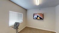 This single-floor fitness room offers a practical and straightforward space dedicated to exercise and wellness. The room is finished with neutral, light beige walls and a textured ceiling, creating a clean and open atmosphere. Durable carpet flooring covers the exercise area, providing a comfortable surface underfoot during workouts. Natural light filters in through multiple windows fitted with horizontal blinds, allowing users to control light levels and maintain privacy while exercising. A wall-mounted flat-screen television is prominently positioned on one wall, enabling users to watch content during their workout sessions. Overhead fluorescent lighting ensures the space remains well-lit at all times. Cardio equipment, including a stationary exercise machine, is present within the room, making it a functional space for fitness routines. The overall design is minimal and utilitarian, prioritizing function over ornamentation. The layout is efficient, making good use of the available footprint while keeping the space uncluttered. This fitness room is well-suited for a residential community or small facility seeking to provide accessible, on-site exercise amenities in a simple, comfortable environment.