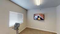 This single-floor fitness room offers a practical and straightforward space dedicated to exercise and wellness. The room is finished with neutral, light beige walls and a textured ceiling, creating a clean and open atmosphere. Durable carpet flooring covers the exercise area, providing a comfortable surface underfoot during workouts. Natural light filters in through multiple windows fitted with horizontal blinds, allowing users to control light levels and maintain privacy while exercising. A wall-mounted flat-screen television is prominently positioned on one wall, enabling users to watch content during their workout sessions. Overhead fluorescent lighting ensures the space remains well-lit at all times. Cardio equipment, including a stationary exercise machine, is present within the room, making it a functional space for fitness routines. The overall design is minimal and utilitarian, prioritizing function over ornamentation. The layout is efficient, making good use of the available footprint while keeping the space uncluttered. This fitness room is well-suited for a residential community or small facility seeking to provide accessible, on-site exercise amenities in a simple, comfortable environment.