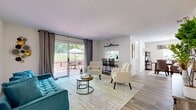 Located at 4577 Williamstown Blvd in Lakeland, Florida, this single-floor residence offers a thoughtfully organized layout encompassing two bedrooms, two bathrooms, a kitchen, a dedicated dining room, and a combined living area. Light-toned wood-look flooring runs throughout the main living spaces, lending a clean, cohesive aesthetic across the home.

The living room serves as the centerpiece of the space, highlighted by large sliding glass doors dressed with floor-length curtains that frame lush green outdoor views and flood the room with natural light. A built-in wall shelf adds a subtle architectural detail. The sliding doors open directly onto a private patio, enclosed by a wood fence and landscaped with small garden beds, offering a comfortable outdoor dining area overlooking an open grassy lawn bordered by mature trees.

The kitchen features a U-shaped layout with ample white cabinetry accented by wood trim, generous counter space, and a window for natural light. Adjacent to the kitchen, the dining room accommodates a full dining set beneath a gold chandelier. Both bedrooms are carpeted and receive natural light from windows. The bathrooms are finished with wood-look flooring, white countertops, and wide vanity mirrors, maintaining the home's consistent, clean design throughout.