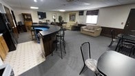 This single-floor space is designed to accommodate a variety of social and recreational activities within one open layout. The room is anchored by a prominent curved bar with a dark countertop and bar-height seating, providing a central hub for socializing. Adjacent to the bar, a pool table adds a recreational element to the space. The flooring transitions between a tiled area near the bar and gray carpeting throughout the main lounge zone, helping to define distinct functional areas.

The lounge area is furnished with multiple sofas and chairs arranged to encourage conversation and relaxation, complemented by side tables, a floor lamp, and decorative touches including framed artwork and potted plants. A wall-mounted flat-screen television is positioned for easy viewing from the seating area. Dark wood wainscoting lines the lower portion of the walls, adding a classic, warm character to the space.

Natural light enters through windows dressed with dark valances, while recessed and flush-mounted ceiling fixtures provide consistent artificial lighting throughout. Additional furniture, including bookcases and storage pieces along the perimeter, contributes to the room's fully equipped, functional atmosphere suited for entertaining and community gatherings.