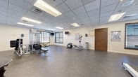 Located on McClellan Loop at Fort Riley, Kansas, this fitness facility offers a thoughtfully organized single-floor layout designed to support a comprehensive workout experience. The space is divided into two distinct areas: a main exercise room and a secondary room, likely used for additional activities or seating.

The main exercise area features durable rubber flooring suited for high-traffic use, providing both safety and comfort during workouts. The room is generously proportioned, allowing for comfortable movement between equipment stations. Cardio machines, including treadmills, an elliptical, and a stationary bike, are positioned throughout the space, while a multi-station strength training unit and additional resistance machines are available for weight training. Wall-mounted televisions provide entertainment during workouts, and informational fitness posters are displayed along the walls.

Natural light enters through multiple windows fitted with horizontal blinds, complementing the bright, recessed fluorescent lighting set within a drop-tile ceiling. Full-length mirrors on one wall enhance the sense of space and allow users to monitor their form. The secondary room features hardwood flooring, a seating area with wooden furniture, and large windows. Together, the two areas create a functional and welcoming environment suited to a range of fitness needs.