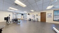 Located on McClellan Loop at Fort Riley, Kansas, this fitness facility offers a thoughtfully organized single-floor layout designed to support a comprehensive workout experience. The space is divided into two distinct areas: a main exercise room and a secondary room, likely used for additional activities or seating.

The main exercise area features durable rubber flooring suited for high-traffic use, providing both safety and comfort during workouts. The room is generously proportioned, allowing for comfortable movement between equipment stations. Cardio machines, including treadmills, an elliptical, and a stationary bike, are positioned throughout the space, while a multi-station strength training unit and additional resistance machines are available for weight training. Wall-mounted televisions provide entertainment during workouts, and informational fitness posters are displayed along the walls.

Natural light enters through multiple windows fitted with horizontal blinds, complementing the bright, recessed fluorescent lighting set within a drop-tile ceiling. Full-length mirrors on one wall enhance the sense of space and allow users to monitor their form. The secondary room features hardwood flooring, a seating area with wooden furniture, and large windows. Together, the two areas create a functional and welcoming environment suited to a range of fitness needs.