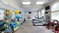 This fitness center offers a thoughtfully designed workout environment that combines functionality with refined architectural details. The space features crown molding and elevated ceilings that lend an upscale feel uncommon in typical gym settings. Flush-mounted square light panels, recessed lighting, and large arched windows along one wall flood the room with ample natural and artificial light, creating a bright and energizing atmosphere.

The cardio zone is well-appointed, housing a row of treadmills and elliptical machines positioned to allow comfortable spacing between users. Wall-mounted flat-screen televisions are strategically placed for viewing during workouts. A floor-to-ceiling mirror runs along one wall, visually expanding the space and providing users with form feedback during exercise.

The functional training area includes a variety of accessories such as stability balls in multiple sizes, resistance bands, and yoga or exercise mats stored on a dedicated rack. The neutral-toned, modular carpet tile flooring adds both comfort underfoot and a polished aesthetic. Overall, the space balances a welcoming, upscale ambiance with practical, comprehensive fitness amenities.