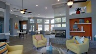 This well-appointed single-floor community lounge offers an open, airy atmosphere anchored by a striking double-sided fireplace clad in mosaic tile, serving as the room's centerpiece. The soaring ceilings are fitted with ceiling fans and recessed lighting, ensuring the space remains bright and comfortable year-round. Large clerestory windows along the upper walls flood the interior with abundant natural light, complementing the cool gray walls and modern finishes throughout.

The layout is thoughtfully divided into distinct functional zones. A casual seating area features plush yellow accent chairs arranged around a glass coffee table, creating an inviting space for relaxed conversation. Adjacent, a more formal lounge area provides additional seating, while a bar-height counter area along one side offers a spot for casual dining or socializing. A secondary seating nook near the windows allows for quieter gatherings with views of the surrounding greenery.

Bold accent walls in warm terracotta add a pop of color and visual contrast to the otherwise neutral palette. The combination of contemporary furnishings, warm lighting, and an open floor plan gives this communal space a welcoming and sophisticated ambiance, well-suited for both everyday lounging and social events.