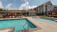 Located at 1410 Nottingham St in Huntsville, Texas, this single-story amenity and clubhouse facility serves as the social and recreational hub of a multi-building residential community. The property is thoughtfully designed to offer a wide range of amenities within a cohesive, well-maintained campus setting.

At the heart of the outdoor space is a resort-style pool area accommodating a substantial number of guests, complemented by a separate wading or spa pool. Surrounding the pools, residents will find an ample sun deck furnished with lounge chairs and chaise lounges, perfect for relaxing outdoors. A pergola-covered outdoor lounge with comfortable seating and grilling stations provides a shaded gathering space, ideal for socializing. Stone-accented columns and iron fencing lend a polished, upscale aesthetic to the pool enclosure.

The interior clubhouse corridor features polished concrete floors, clean white walls, and recessed lighting, projecting a modern and well-kept atmosphere. Directional signage points residents toward key amenities including a dining room, fitness center, game lounge, and member lounge — indicating a robust offering of indoor communal spaces.

The surrounding grounds are neatly landscaped with manicured lawns and greenery, complementing the blue-gray siding exterior of the multi-story residential buildings. The overall ambiance of the property is welcoming, active, and community-oriented.