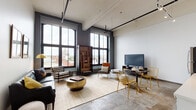 This urban loft showcases a striking industrial aesthetic defined by soaring ceilings, exposed ductwork, and original concrete floors that run throughout the space. The open-concept living and dining area is a standout, anchored by dramatic floor-to-ceiling black-framed windows that flood the interior with abundant natural light and frame sweeping city rooftop views. The living area features a tufted leather sofa, plaid accent chair, brass coffee table, and a warm wood open-shelving unit, creating a sophisticated yet inviting atmosphere. A dedicated dining nook with wire-frame gold chairs adds a modern industrial touch. The kitchen is outfitted with stainless steel appliances, white cabinetry, a grey tile backsplash, and in-unit laundry, balancing functionality with clean contemporary design. The bedroom offers a quiet retreat with neutral walls, mixed-media flooring, and ample room for furnishings. The bathroom features a white vanity with a large mirror, mosaic tile accents, and a shower-tub combination. Exposed structural columns and ceiling-mounted track lighting add to the authentic industrial character found throughout this thoughtfully designed urban residence.