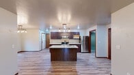 This single-story home showcases a cohesive, modern design unified by warm-toned luxury vinyl plank flooring and rich dark wood trim that flows throughout the interior. The open-concept kitchen serves as the heart of the home, anchored by a central island with a granite-look countertop and integrated sink. Dark espresso cabinetry lines the walls, complemented by stainless steel appliances including a refrigerator and range with overhead ventilation. An industrial-style Edison bulb chandelier suspended from a wood-and-pipe fixture adds a distinctive design element above the dining area, which also benefits from a window that brings in natural light. The kitchen opens directly into a spacious living area, creating a seamless flow ideal for everyday living and entertaining. Additional rooms offer flexible space, each finished with the same plank flooring and neutral wall tones, providing a clean, move-in-ready canvas. The full bathroom features a dark wood vanity with a stone-look countertop, a wall-mounted mirror with bar lighting, and a tub-shower combination. The attached garage provides generous storage and parking space, finished with a concrete floor and an automated door opener.