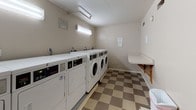 The laundry room in Building 380 is a single-floor shared facility designed to serve the needs of residents with practicality and efficiency. The room is equipped with a row of front-loading washing machines and top-loading dryers lined along one wall, offering multiple units to accommodate simultaneous use. Overhead fluorescent lighting illuminates the space evenly, ensuring good visibility throughout. A built-in folding counter runs along the opposite wall, providing a dedicated surface for sorting and folding laundry. The floor features a classic checkered vinyl tile pattern in muted tan and brown tones, contributing to a clean and functional aesthetic. The walls are finished with a textured plaster surface in a neutral off-white tone. A window and a doorway on one end of the room connect directly to the adjacent covered parking structure, allowing for convenient access and a degree of natural light. Informational notices are posted on the wall, offering usage guidelines for residents. The layout is straightforward and well-organized, maximizing the usable floor space for laundry activities. A fire extinguisher is mounted near the entrance, reflecting attention to safety within the facility.