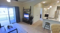 This single-floor residence features an open-concept layout that seamlessly connects the living area to the kitchen. The carpeted living room is finished with crown molding and provides a comfortable, functional space centered around a ladder-style media shelf holding a flat-screen television. A decorative area rug adds texture underfoot, while a potted plant introduces a touch of greenery. Patterned curtains frame a sliding glass door that fills the room with natural light and provides access to an outdoor area, offering views beyond the unit. A ceiling fan enhances comfort overhead. The kitchen is visible from the living space and features dark cabinetry, granite or stone-look countertops, and a raised breakfast bar with seating. The kitchen is well-lit and includes a sink and ample counter space, making it both practical and visually appealing. A hallway leads toward additional rooms, with a closet door visible along the corridor. Overall, the space presents a clean, neutral palette with warm tones, creating a cohesive and inviting atmosphere throughout.