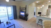 This single-floor residence features an open-concept layout that seamlessly connects the living area to the kitchen. The carpeted living room is finished with crown molding and provides a comfortable, functional space centered around a ladder-style media shelf holding a flat-screen television. A decorative area rug adds texture underfoot, while a potted plant introduces a touch of greenery. Patterned curtains frame a sliding glass door that fills the room with natural light and provides access to an outdoor area, offering views beyond the unit. A ceiling fan enhances comfort overhead. The kitchen is visible from the living space and features dark cabinetry, granite or stone-look countertops, and a raised breakfast bar with seating. The kitchen is well-lit and includes a sink and ample counter space, making it both practical and visually appealing. A hallway leads toward additional rooms, with a closet door visible along the corridor. Overall, the space presents a clean, neutral palette with warm tones, creating a cohesive and inviting atmosphere throughout.