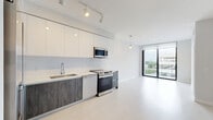 Located at 150 S Bryan Rd in Dania Beach, Florida, this modern residence offers a thoughtfully designed one-bedroom, two-bathroom layout across a single floor. The open-concept living and kitchen area is anchored by a sleek, contemporary kitchen featuring two-tone cabinetry — warm wood-grain lower cabinets paired with crisp white upper cabinets — complemented by white quartz countertops, a white subway tile backsplash, and a full suite of stainless steel appliances including a French door refrigerator, electric range, and over-the-range microwave. Light wood-toned flooring flows throughout, enhancing the bright, airy atmosphere. The living area opens to large floor-to-ceiling sliding glass doors leading to a balcony, flooding the space with natural light and providing outdoor views. The bedroom is a calm, neutral retreat with a large window framing views of the surrounding community and ample room for furnishings. The two bathrooms are finished in a clean, modern style, one featuring a soaking tub and shower combination with light stone-look tile surround, a floating wood vanity, and contemporary fixtures. Track lighting and pendant accents throughout add a polished, functional touch to this well-appointed Dania Beach residence.