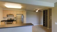 This single-floor space showcases an open-concept layout that seamlessly connects the kitchen and living area. The kitchen is equipped with stainless steel appliances, including a refrigerator and electric range, complemented by light wood cabinetry that extends to the ceiling for generous storage. Dark granite-style countertops provide a contrasting accent, while a peninsula breakfast bar serves as a natural divider between the kitchen and the adjoining living space. Overhead, a flush-mount fluorescent fixture illuminates the kitchen, while a ceiling fan with a light kit serves the living area, promoting air circulation and ambient lighting. The living space features neutral gray walls, carpeted flooring, and a built-in shelving unit along one wall, offering both functional storage and display space. The overall design is practical and cohesive, with a neutral color palette of grays and beiges creating a calm, understated ambiance throughout. The open floor plan enhances the sense of space, making the layout feel connected and versatile for everyday living.