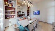 This thoughtfully designed dining space combines contemporary and transitional elements to create a warm yet sophisticated atmosphere. At its center sits a substantial concrete-top dining table paired with upholstered chairs in neutral tones and accented by teal seating, lending a pop of color to the refined palette. A striking cluster of hand-blown glass pendant lights suspended at varying heights from a black rail serves as the room's dramatic focal point, casting a warm, ambient glow over the dining area. One wall features a custom built-in unit with deep burgundy-backed open shelving that frames a large mirror, creating depth and visual interest while providing display space for decorative objects. A built-in upholstered banquette beneath the shelving is dressed with an eclectic mix of patterned and coral accent pillows, adding comfort and a vibrant energy. Light hardwood flooring runs throughout, complementing the neutral wall tones. A wall-mounted flat-screen television adds a modern, functional element. Elegant black-framed glass French doors connect the dining room to adjacent spaces and an outdoor area, inviting abundant natural light and offering glimpses of the exterior. A colorful abstract artwork and a classic gray sideboard complete the room's curated, art-forward aesthetic.