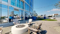 This expansive rooftop terrace is a thoughtfully designed outdoor amenity space set against the backdrop of a modern glass-curtain high-rise building. The terrace is organized into several distinct functional zones, each offering a unique experience. At its centerpiece is a generously sized circular artificial turf lawn, ideal for lawn games such as cornhole and bocce ball, with string lights overhead adding a festive ambiance during evening hours. Flanking the lawn are bold, oversized cobalt-blue planters housing mature trees that provide both shade and striking visual interest. A dedicated lounge area features a round concrete fire pit surrounded by Adirondack chairs, creating an inviting space for relaxed gatherings. A large outdoor dining table with wicker chairs accommodates group dining and social events. The terrace is finished with smooth concrete pavers throughout, and low perimeter walls with partial glass railings allow for unobstructed panoramic views of the surrounding urban skyline, including a notable historic tower and palm-tree-lined streets. The full-height glass facade of the adjacent building reflects the open sky, further amplifying the sense of light and openness that defines this exceptional outdoor space.
