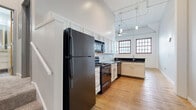 Located at 1278 W Ninth St in Cleveland's vibrant Ohio City neighborhood, this 2-bedroom, 2-bathroom unit showcases industrial loft character throughout. Soaring ceilings with exposed ductwork, conduit, and pendant lighting set the tone from entry, where warm hardwood floors extend through the hallway, living area, and kitchen. The open kitchen is well-appointed with white shaker cabinetry, dark granite countertops, a classic subway tile backsplash, and black stainless appliances including a gas range, built-in microwave, and dishwasher. Multi-pane windows with bold red frames flood the kitchen and living areas with natural light and add an industrial-chic accent. A breakfast bar provides casual dining space adjacent to the kitchen. The living area benefits from the same soaring ceiling height and exposed white brick, adding texture and historic character. Both bedrooms feature plush carpeting and generous closet space, with interior windows that borrow light from the main living areas. The two bathrooms are finished with granite-topped vanities and tile flooring; one includes a glass-enclosed walk-in shower. Throughout, the unit blends historic architectural details with modern finishes, creating a distinctive and functional urban living space.