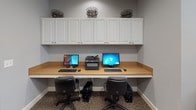 This single-floor business center is thoughtfully designed to support productivity and comfort. The space is divided into two distinct functional zones, connected by a white six-panel door. The walls throughout are finished in a soft blue-gray tone, complemented by white baseboards and trim, creating a calm and professional atmosphere.

The first zone features a dedicated computer station with a long, built-in wood-tone countertop desk accommodating two desktop computers and a multifunction printer. Above the desk, a row of white raised-panel cabinetry provides ample closed storage. Ergonomic task chairs and carpeted flooring complete this workstation area.

The adjacent zone serves as a collaborative workspace, featuring an L-shaped built-in desk with a warm wood-tone surface that wraps along two walls, offering generous counter space for multiple users. Upholstered chairs with nailhead trim line the desk, providing comfortable seating. A large window with white horizontal blinds fills the room with natural light, while a framed abstract canvas in neutral earth tones adds a decorative focal point. Recessed ceiling lighting and a HVAC vent ensure a comfortable environment. The tile flooring in the connecting corridor transitions to plush carpet within the workspace, enhancing the overall sense of warmth and professionalism.