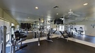 This fitness center is a thoughtfully designed workout facility that provides a comprehensive environment for both cardio and strength training. The space is lined with floor-to-ceiling mirrors along the walls, which enhance the sense of openness and allow exercisers to monitor their form throughout their routines. A row of Matrix-brand treadmills and elliptical machines lines one side of the room, offering ample cardio options, while a dedicated strength training area with cable machines and weight benches occupies the opposite end. The flooring features durable, cushioned rubber matting throughout, providing both safety and comfort underfoot. Recessed lighting is complemented by elegant ceiling-mounted light fixtures and multiple ceiling fans, which help maintain air circulation and a comfortable workout environment. Large windows along the far wall allow natural light to pour into the space, creating a bright and energizing atmosphere. The overall ambiance is clean, functional, and well-maintained, making it a practical and inviting space suited for a full range of fitness activities.