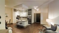 This single-floor residence offers a thoughtfully designed open-concept layout that seamlessly connects the kitchen and living areas. The kitchen is a standout feature, showcasing white cabinetry that extends to the upper walls with open display shelving, a mosaic tile backsplash, and a full suite of stainless steel appliances including a refrigerator with bottom freezer and a stainless range with microwave. A central island with a light stone countertop provides additional prep space and casual seating with industrial-style bar stools, making it ideal for both cooking and entertaining. Pendant lights hang above the island, complementing the flush-mount ceiling fixtures that provide warm, even illumination throughout the space. Rich, dark hardwood-style flooring runs continuously through the kitchen, dining, and living areas, adding warmth and cohesion to the open floor plan. The living area is generously proportioned, flowing naturally from the kitchen. A dedicated wine rack column adds a refined architectural detail between the kitchen and the entry corridor. The entry features a bold, dark-paneled door that creates a striking contrast against the neutral, light-toned walls. The overall ambiance is modern yet welcoming, combining clean lines with warm finishes for a comfortable and functional living environment.