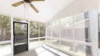 Located at 7301 Brainerd Rd in Chattanooga, Tennessee, this single-floor screened porch offers a serene and light-filled retreat with direct views of a verdant, tree-lined backyard. The space is enclosed by floor-to-ceiling screened window panels on three sides, allowing natural light to pour in from nearly every angle while maintaining a comfortable, open-air feel. A vaulted ceiling adds a sense of height and spaciousness, further enhancing the airy atmosphere. A ceiling fan is centrally mounted overhead, providing efficient air circulation throughout the warmer months. The concrete floor is clean and neutral, complementing the crisp white walls and bright white window framing. A black-framed door provides direct access to the backyard, connecting the interior to the outdoor green space effortlessly. The surrounding mature trees are clearly visible through the screens, offering a natural, private backdrop and a peaceful ambiance. This screened porch is well-suited for relaxing, entertaining, or simply enjoying the outdoors in a sheltered environment.