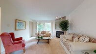 Located at 1011 East Sharpsburg Avenue in Spokane, Washington, this single-floor residence offers a well-organized layout with distinct living spaces throughout. The spacious living room serves as the heart of the home, featuring a tiled fireplace flanked by a large mirror above the mantle, soft carpet flooring, and generous windows that flood the room with natural light and frame views of surrounding greenery. A sliding glass door leads to a private balcony overlooking mature trees, offering a serene outdoor retreat. The dining area is bright and airy, with windows providing additional garden views and ceiling fan with recessed lighting overhead, accommodating a full dining set comfortably. The kitchen is efficiently designed with light wood cabinetry, ample counter space, a full suite of white appliances including a refrigerator, electric range, and a double sink. The bedroom is generously proportioned with closet storage and connects conveniently to adjoining spaces. The bathroom features a single-sink vanity with light wood cabinetry, a mirror with overhead lighting, tile flooring, and a walk-in shower enclosure. Throughout the residence, neutral tones, plush carpeting, and an abundance of natural light from tree-lined surroundings create a calm and comfortable living environment.