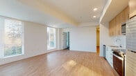 This single-floor residence is designed with a clean, contemporary aesthetic defined by crisp white walls, light hardwood-style flooring, and an abundance of natural light. The open-concept living and dining area is generously proportioned, with large windows fitted with horizontal blinds that allow soft, diffused light to fill the space. Recessed lighting complements the natural illumination, and a tray ceiling detail adds subtle architectural interest.

The kitchen is efficiently laid out along one wall, featuring warm natural wood upper cabinetry paired with white lower cabinets and light countertops. A white subway tile backsplash, undermount sink, and a full suite of stainless steel appliances — including a refrigerator, electric range, built-in microwave, and dishwasher — complete the functional and stylish space.

Bedrooms are carpeted in neutral tones, offering a comfortable contrast to the hard flooring in the main living areas. At least one bedroom includes a sliding-door closet. A hallway connects the rooms, maintaining a logical and practical flow throughout.

The bathroom is finished in an all-white palette, featuring a soaking tub with tiled surround, chrome fixtures, a white vanity with an integrated sink, and light hardwood-style flooring, contributing to the unit's cohesive modern feel.