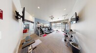 Located at 10602 Burns Rd in Pasco, WA, this single-floor fitness center offers a well-equipped and thoughtfully designed workout space. The facility spans a generous footprint, providing ample room for a diverse range of exercise equipment and activities. The interior is finished with durable patterned carpet tiles accented by red fitness mats, white walls, and blue accent walls that create a clean, modern aesthetic. Recessed lighting illuminates the space evenly throughout.

A standout architectural feature is the large, full-width garage-style glass door on one wall, flooding the workout area with abundant natural light and offering open views of the surrounding community. This transparency creates an airy, inviting atmosphere that enhances the exercise experience.

The cardio zone is well-stocked with multiple treadmills, elliptical machines, and a stair climber, all arranged to take advantage of the natural light from the glass door. A dedicated strength training area features a multi-functional cable machine, adjustable benches, and a full rack of dumbbells in varying weights. Wall-mounted flat-screen televisions are positioned for convenient viewing during workouts.

An adjacent lounge area, visible through glass sliding doors, features plush seating with red accent chairs, hardwood-style flooring, and soaring ceilings with tall windows — offering a welcoming space for residents to relax before or after their workout.