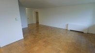 This single-floor space presents a straightforward and functional layout with a clean, neutral aesthetic throughout. The most notable design feature is the classic parquet wood flooring, which covers the main living area in a warm, geometric pattern that adds visual texture and timeless character to the space. The walls are finished in crisp white, contributing to a bright and open atmosphere that makes the interior feel airy and expansive. The open-plan living area provides generous room for a variety of furniture arrangements, offering flexibility for different uses. A doorway leads to an adjoining area, where built-in shelving provides convenient storage or display space — a practical and permanent architectural detail. A baseboard heating unit is visible along one wall, indicating a reliable in-unit heating system. The overall layout is efficient and uncluttered, with clean sightlines across the main room. The combination of parquet flooring, white walls, and open space gives the interior a classic, understated quality that serves as a neutral canvas, well-suited to a range of interior styles and personal touches.
