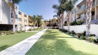 This residential community is defined by its thoughtfully designed outdoor spaces and clean architectural presence. The grounds feature expansive, manicured lawns that stretch between multi-story buildings, creating an open and park-like atmosphere throughout the property. Wide concrete walkways wind through the courtyards, flanked by neatly trimmed grass and carefully tended garden beds filled with drought-tolerant shrubs, flowering plants, and ornamental grasses.

Tall, mature palm trees are planted throughout the common areas, lending a warm, Southern California character to the environment. The buildings themselves present a neutral stucco exterior with accents of yellow, giving the community a bright and cohesive aesthetic. Balconies are visible along the upper floors, offering residents outdoor access and views of the greenery below.

A covered lounge area near the entrance provides a shaded retreat, furnished with a classic wooden bench and surrounded by potted tropical plants. This sheltered space looks out over the green courtyard and adjacent buildings, offering a comfortable spot to relax and enjoy the surroundings. The overall ambiance of the community is serene, open, and well-kept, making the outdoor common areas a central and welcoming feature of the property.