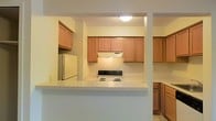 This single-floor residence offers a practical and well-organized layout designed for comfortable everyday living. The kitchen serves as a central highlight of the space, featuring warm honey-toned wood cabinetry that extends across multiple walls, providing generous storage for all household needs. Upper and lower cabinets are thoughtfully arranged to maximize functionality, while a raised peninsula counter defines the kitchen boundary and creates a natural division between the cooking area and the adjoining space. The kitchen is equipped with a white coil-top electric range with an overhead ventilation hood, as well as a full-size white refrigerator positioned neatly against the wall. Light-colored countertops complement the cabinetry and contribute to a clean, cohesive aesthetic throughout the kitchen. The walls are finished in a soft, neutral tone that keeps the space feeling bright and open. Overhead flush-mount lighting provides consistent illumination across the kitchen area. Light-toned flooring ties the space together, adding to the overall sense of cleanliness and simplicity. The open sightlines between the kitchen and adjacent areas enhance the feeling of connectedness throughout the single-floor layout, making the space both efficient and easy to navigate.