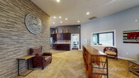 Located at 5800 S Jefferson Commons Cir in Kalamazoo, MI, this single-floor community clubhouse offers a well-appointed gathering space designed for both relaxation and productivity. The entry foyer makes an immediate impression with its striking stacked-stone accent wall, decorative oversized clock, and warm wood-look flooring, setting an inviting tone throughout.

The main lounge is a generously sized open area featuring hardwood-style flooring, vaulted ceilings with recessed lighting, and a bold stone fireplace wall with a mounted flat-screen TV. Comfortable leather seating, a vibrant area rug, and a full-size pool table beneath a statement chandelier make this an ideal space for socializing and recreation. Large French doors along one wall invite natural light and provide access to an outdoor area.

Adjacent to the lounge, the kitchenette is equipped with dark espresso cabinetry, mosaic tile backsplash, light countertops, and a stainless steel refrigerator — ideal for hosting events or casual gatherings.

A dedicated reception and seating area features a herringbone-patterned floor, large windows with ample natural light, and a variety of seating arrangements suitable for casual meetings or quiet relaxation. A private conference room with a glass door offers a functional workspace for meetings and collaboration. Throughout the space, cool gray walls and carefully selected décor contribute to a modern yet comfortable community atmosphere.