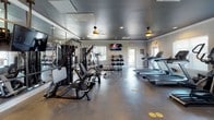 This single-floor fitness center in Lebanon, Tennessee, is thoughtfully organized into distinct workout zones, each designed to support a comprehensive exercise experience. The main strength and cardio area features durable epoxy flooring, a dark-painted ceiling with recessed lighting and ceiling fans, and crown molding along light gray walls. Full-length mirrored panels line one wall, visually expanding the space while allowing users to monitor their form. This primary zone is equipped with a multi-station cable machine, adjustable benches, a dumbbell rack, and multiple treadmills positioned near windows that allow natural light to filter in. A mounted flat-screen television adds convenience for longer cardio sessions. A dedicated cardio room, finished with neutral carpeting and a dark ceiling, houses elliptical machines and features a pass-through window offering views into the main floor area. A separate cycling studio offers a more intimate setting, with indoor cycling bikes, TRX suspension training straps mounted to a wall-mounted rig, and an accent wall of reclaimed wood-style paneling providing a distinct aesthetic. The space is rounded out by direct exterior door access, ensuring easy entry and exit throughout the facility.