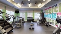 This fitness center is a comprehensive, single-floor facility designed to support a full spectrum of workout routines. The space is thoughtfully laid out, with distinct zones dedicated to cardio training, strength equipment, and functional fitness. The cardio area features a lineup of treadmills, elliptical machines, and a StairMaster, all positioned in front of windows fitted with white horizontal blinds that allow ample natural light to fill the room. Ceiling fans with integrated lighting fixtures are mounted on a recessed tray ceiling, contributing to both comfort and visual appeal. The strength and free weights section includes a well-stocked dumbbell rack, a weight bench, and a cable machine, providing options for a thorough resistance workout. Stability and medicine balls in various sizes are neatly organized on a dedicated rack, supporting functional and core training. Recumbent bikes offer lower-impact cardio options along one wall. A vibrant, large-format word-cloud mural spanning fitness-related terms — including "exercise," "lifestyle," "energy," and "motivation" — energizes the space with an inspirational visual backdrop. Wall-mounted flat-screen televisions are positioned throughout for entertainment during workouts. A full-length mirror expands the sense of space, while durable rubber flooring ensures safety and functionality throughout.