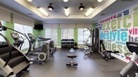 This fitness center is a comprehensive, single-floor facility designed to support a full spectrum of workout routines. The space is thoughtfully laid out, with distinct zones dedicated to cardio training, strength equipment, and functional fitness. The cardio area features a lineup of treadmills, elliptical machines, and a StairMaster, all positioned in front of windows fitted with white horizontal blinds that allow ample natural light to fill the room. Ceiling fans with integrated lighting fixtures are mounted on a recessed tray ceiling, contributing to both comfort and visual appeal. The strength and free weights section includes a well-stocked dumbbell rack, a weight bench, and a cable machine, providing options for a thorough resistance workout. Stability and medicine balls in various sizes are neatly organized on a dedicated rack, supporting functional and core training. Recumbent bikes offer lower-impact cardio options along one wall. A vibrant, large-format word-cloud mural spanning fitness-related terms — including "exercise," "lifestyle," "energy," and "motivation" — energizes the space with an inspirational visual backdrop. Wall-mounted flat-screen televisions are positioned throughout for entertainment during workouts. A full-length mirror expands the sense of space, while durable rubber flooring ensures safety and functionality throughout.