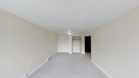 This single-floor space offers a straightforward and functional layout designed for comfortable everyday living. The main room is generously proportioned, featuring soft wall-to-wall carpeting in a neutral gray tone that complements the warm, cream-colored walls and crisp white ceiling. A flush-mount ceiling light provides overhead illumination, keeping the space bright and welcoming. A built-in closet with open shelving provides convenient storage, while a separate entry door adds a sense of defined transition between interior spaces. Baseboard heating runs along the perimeter of the room, ensuring efficient and even warmth throughout. The clean lines, minimal detailing, and consistent neutral tones create a calm and versatile environment that can readily accommodate a variety of furniture arrangements and personal styles. The overall ambiance is understated and practical, offering a blank canvas that is easy to personalize. The space is well-maintained, with smooth walls and clean finishes throughout, making it a functional and move-in ready environment.