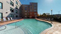 Located at 401 S Main St in downtown Akron, Ohio, this facility showcases a well-appointed outdoor pool area and supporting amenities within a contemporary multi-story building. The outdoor pool is generously sized with a freeform shape, surrounded by an expansive pool deck lined with lounge chairs and umbrellas, ideal for relaxation and recreation. The building's exterior combines brick and modern paneling, rising several stories and framing the pool area with a clean, urban aesthetic.

Inside, the ground-floor common areas feature light-colored large-format tile flooring that lends a bright, clean feel throughout. A dedicated restroom corridor provides access to separate men's and women's facilities, both ADA-accessible and clearly marked with signage. The restroom anteroom is finished with neutral walls and tile floors, maintaining a functional and tidy appearance.

A notable interior feature includes dual stainless steel drinking fountains — one at standard height and one at ADA-accessible height — mounted against a striking dark tile accent wall. This area transitions directly to glazed entry doors that open to the outdoor pool deck, providing a seamless connection between the indoor and outdoor spaces. Natural light filters in through the glass doors, brightening the corridor and offering views of the poolside seating area beyond.