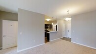 This single-floor residence offers a thoughtfully designed open-concept layout that seamlessly connects the living and kitchen areas, creating a spacious and cohesive environment. The living area features plush carpeted flooring and soft neutral wall tones that contribute to a warm, welcoming ambiance throughout. An elegant pendant light fixture hangs overhead, adding a decorative touch to the space.

The kitchen is positioned adjacent to the living area and is defined by a peninsula-style breakfast bar that acts as both a functional workspace and a natural divider between the two spaces. Dark espresso cabinetry provides a striking contrast against the light countertops, while stainless steel appliances — including a microwave and range — lend a modern, polished feel. Additional pendant lighting above the kitchen area enhances both task visibility and visual appeal.

A tiled entryway near the front door provides a practical transition zone from the exterior into the living space. The open sightlines from the living area through to the kitchen and entry corridor give the residence a sense of depth and flow. Overall, the space balances modern finishes with a neutral, versatile palette, making it a functional and aesthetically cohesive home.