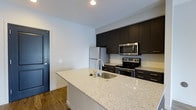 Located at 3443 Park Street in Grove City, Ohio, this single-floor space presents a clean, modern aesthetic throughout. The kitchen serves as a standout feature, showcasing a large island with a light granite countertop and an integrated sink, offering ample workspace and a functional layout ideal for cooking and entertaining. Dark espresso-toned cabinetry lines the walls from counter to ceiling, providing generous storage while creating a striking visual contrast against the light countertops and white tile backsplash. Stainless steel appliances, including a refrigerator, electric range, and over-the-range microwave, complete the kitchen's contemporary look. Warm hardwood-style flooring flows throughout, adding depth and cohesion to the space. The walls are finished in a crisp, neutral white, enhancing the sense of brightness and openness. Recessed lighting provides clean, even illumination across the kitchen area. A bold navy-painted entry door adds a thoughtful design accent, offering a pop of color against the otherwise neutral palette. Overall, the space combines modern finishes with a practical, well-organized layout, creating an inviting and functional environment.