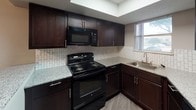 This single-floor unit offers a well-organized layout with consistent design elements throughout. Dark hardwood-style flooring flows seamlessly across the living areas and bedrooms, complemented by neutral taupe-toned walls and crisp white baseboards that create a clean, cohesive aesthetic. Textured ceilings are found throughout the unit.

The living area is generously proportioned, featuring a ceiling fan with integrated lighting and a sliding glass door dressed with vertical blinds, allowing abundant natural light to fill the space while providing direct access to an outdoor balcony or patio area.

The kitchen is a functional highlight, equipped with rich dark espresso cabinetry, granite-look countertops, a classic tile backsplash, and black appliances including an electric range, over-the-range microwave, and a stainless steel sink. A window above the sink provides additional natural light.

The bedroom spaces are well-sized and open, maintaining the same warm flooring and neutral wall palette. Ceiling fans are present, and vertical blinds on windows offer light control and privacy. Additional rooms offer access to closet spaces for storage.

The bathroom includes a full tub and shower combination with white tile surround, a toilet, and built-in open shelving for added storage convenience. Overall, the unit presents a practical and comfortable living environment with thoughtful finishes throughout.
