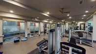 Located at 5225 Las Colinas Blvd in Irving, Texas, this fitness center offers a well-appointed, single-floor workout facility designed to support a full range of exercise needs. The space is thoughtfully divided into distinct functional zones, seamlessly connected by an open layout and clean, contemporary finishes throughout.

The cardio area features multiple treadmills and additional cardio machines positioned beneath soaring double-height windows that flood the space with natural light and provide views of the surrounding outdoor courtyard. Warm hardwood-style flooring grounds this zone, contributing to an inviting atmosphere.

The strength training area is thoroughly equipped with Life Fitness selectorized cable machines, a Smith machine with bench press, and a full dumbbell rack, all set against floor-to-ceiling mirrored walls that enhance the sense of openness and allow users to monitor form. Recessed lighting and ceiling fans maintain a comfortable environment throughout.

A dedicated functional training zone includes a balance disc and additional cable systems, while glass-paneled sliding doors separate private areas, likely housing changing or sauna facilities. Secure keycard-access entry points are visible at multiple entryways, ensuring controlled access. The polished, neutral color palette of gray, white, and blue accents creates a professional and motivating atmosphere suited for daily use.