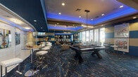 This single-floor entertainment lounge in North Little Rock, Arkansas, is a thoughtfully designed social space that combines upscale aesthetics with a wide range of recreational amenities. The room is defined by its striking interior design, featuring bold teal and cream horizontal striped walls, a deep blue coffered ceiling accented with ambient LED lighting, and a richly patterned dark blue carpet with gold marbling throughout.

At the center of the space sits a full-size pool table, positioned beneath elegant chandelier pendant lighting that adds a refined touch to the room. Surrounding the table is a generous lounge area furnished with neutral-toned armchairs and sofas arranged around circular wood accent tables, creating comfortable seating clusters for socializing. Vibrant, large-scale pop-art portraits adorn the walls, contributing to the space's energetic, contemporary atmosphere.

Toward the rear of the room, a dedicated bar area features bar-height counters and stools, ideal for entertaining. A separate high-top table zone provides additional gathering space. Floor-to-ceiling glass panels and large windows along one wall allow natural light to fill the room and provide views of the outdoor area beyond. A wall-mounted flat-screen television and a popcorn machine add further entertainment options, making this lounge a well-rounded, versatile venue.
