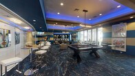 This single-floor entertainment lounge in North Little Rock, Arkansas, is a thoughtfully designed social space that combines upscale aesthetics with a wide range of recreational amenities. The room is defined by its striking interior design, featuring bold teal and cream horizontal striped walls, a deep blue coffered ceiling accented with ambient LED lighting, and a richly patterned dark blue carpet with gold marbling throughout.

At the center of the space sits a full-size pool table, positioned beneath elegant chandelier pendant lighting that adds a refined touch to the room. Surrounding the table is a generous lounge area furnished with neutral-toned armchairs and sofas arranged around circular wood accent tables, creating comfortable seating clusters for socializing. Vibrant, large-scale pop-art portraits adorn the walls, contributing to the space's energetic, contemporary atmosphere.

Toward the rear of the room, a dedicated bar area features bar-height counters and stools, ideal for entertaining. A separate high-top table zone provides additional gathering space. Floor-to-ceiling glass panels and large windows along one wall allow natural light to fill the room and provide views of the outdoor area beyond. A wall-mounted flat-screen television and a popcorn machine add further entertainment options, making this lounge a well-rounded, versatile venue.
