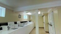 This single-floor shared laundry facility is designed with practicality and efficiency in mind. The room features a row of top-loading washing machines lined along one wall, providing ample capacity for multiple users. Complementing the washers, a set of dryers is positioned along the adjacent section of the room, ensuring a complete and convenient laundry experience. A central support column divides the space naturally, while maintaining an open and accessible flow throughout. The walls are finished in a soft, neutral tone that contributes to a clean and welcoming atmosphere. Overhead, flush-mounted fluorescent light fixtures provide bright, even illumination across the entire room. A small window along one wall allows for a modest amount of natural light to supplement the interior lighting. The flooring is light-colored and easy to maintain, reinforcing the overall hygienic and orderly environment. A framed notice board is mounted on the wall, offering a practical space for posting guidelines or announcements. The facility is thoughtfully organized to accommodate simultaneous use, making it a functional and well-maintained amenity for its users.