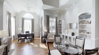 This single-floor space showcases an open, flowing layout that seamlessly connects multiple living and dining areas, creating a sense of generous scale and versatility. The architecture is immediately striking, with soaring vaulted ceilings in the main living zone transitioning to a recessed tray ceiling in the adjacent dining area, adding visual depth and dimension throughout.

Natural light is a defining characteristic of this space. A grand arched window dominates one wall, flooding the interior with bright, diffused sunlight that reflects beautifully off the warm hardwood floors. Additional windows dressed in elegant patterned drapery further enhance the airy atmosphere, while plantation shutters offer light control and privacy.

The space features two distinct dining areas — one anchored by a round glass-top table with upholstered chairs, and another with a dark wood dining set — providing flexible entertaining options. A built-in white cabinetry unit with glass-front display cases and open shelving offers both storage and a refined decorative focal point. A cozy armchair and a console table with a floor lamp define a relaxed transitional seating nook. The neutral white and grey palette, recessed lighting, and ceiling fan complete the sophisticated, welcoming ambiance.