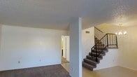 This two-story residence offers a straightforward and functional layout spread across two levels. The main floor features a spacious open living area finished with carpet flooring, neutral white walls, and a textured ceiling that adds subtle visual character throughout the space. A central structural column anchors the layout, while a hallway leads toward additional rooms, including what appears to be a bathroom with warm interior lighting visible in the background.

A carpeted staircase with a sleek dark metal railing connects the main floor to the upper level, serving as a practical and visually clean architectural element within the home. The staircase is positioned prominently near the entry area, making the transition between floors both accessible and straightforward.

The overall color palette is light and neutral, with white walls and ceilings contributing to a bright, open feel throughout the main living area. HVAC venting is integrated into the walls, reflecting a practical approach to the home's infrastructure. The combination of carpet in the living and stair areas with hard flooring at the base entry creates a clear distinction between zones, adding to the home's organized and livable character. This space presents a solid foundation well-suited for comfortable everyday living.
