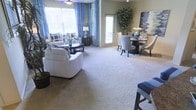This single-floor space showcases an open-concept layout that seamlessly integrates a living room and dining area into one cohesive environment. The living area is furnished with plush white seating, a dark oval coffee table, and decorative blue-and-white throw pillows, creating a clean, coordinated aesthetic. Blue curtain panels frame multiple windows, allowing generous amounts of natural light to fill the room while adding a pop of color against the neutral walls.

The flooring throughout is wall-to-wall carpet in a warm beige tone, contributing to a comfortable and inviting atmosphere. A ceiling fan is centrally positioned overhead, enhancing both function and airflow within the space.

The adjacent dining area features a round dark table surrounded by elegantly styled upholstered chairs, complemented by a tall indoor potted tree that adds a natural, organic element to the space. A bold abstract wall canvas in slate and blue tones serves as a striking focal point behind the dining arrangement.

Overall, the space presents a well-balanced blend of comfort and style, with consistent blue and neutral tones tying the living and dining zones together in a harmonious, well-lit environment.